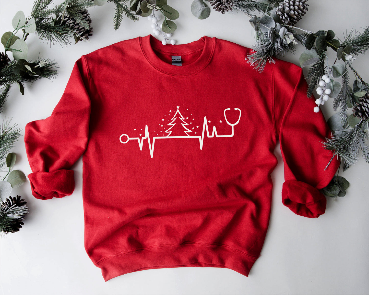 A red sweater with a white Christmas tree and stethoscope design, surrounded by pine branches and greenery.