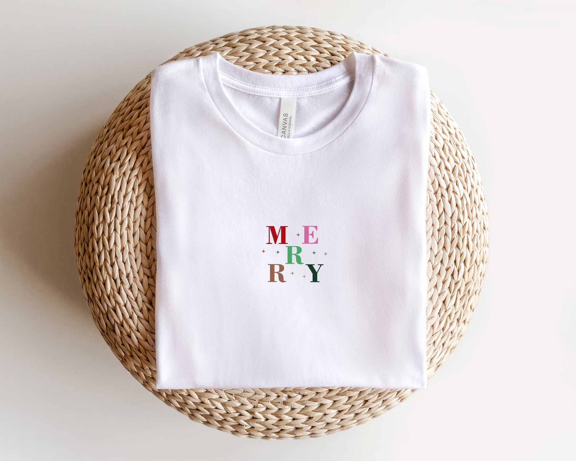 A white t-shirt with the word "MERRY" printed on it, laid out on a woven basket or mat.