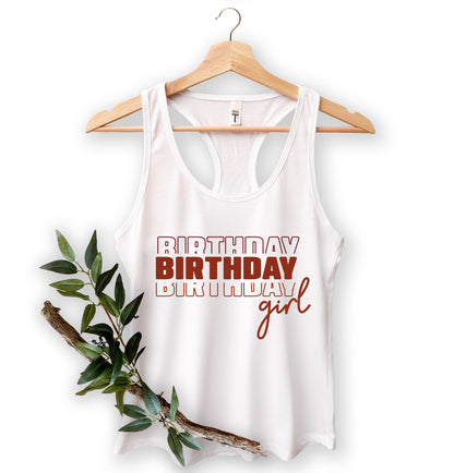 A white tank top with the text "BIRTHDAY BIRTHDAY girl" printed on it, hanging on a wooden hanger.
