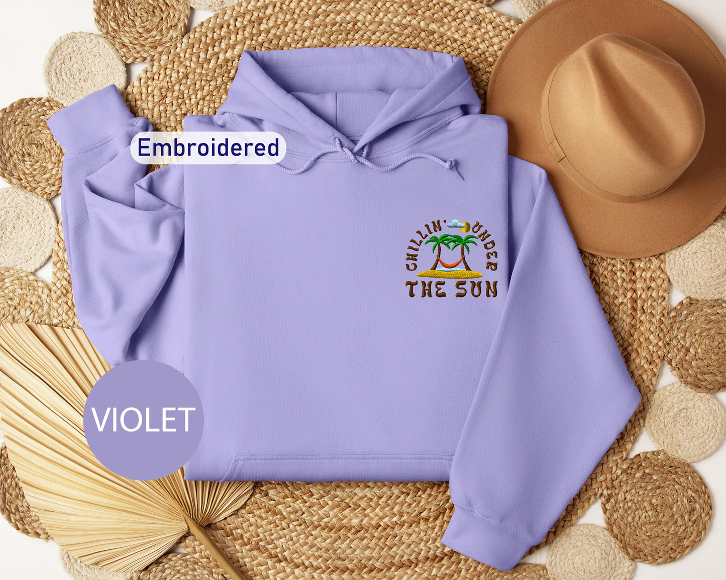 a purple hoodie with a tropical beach design, a brown hat, and a woven straw hat, all arranged on a woven mat.