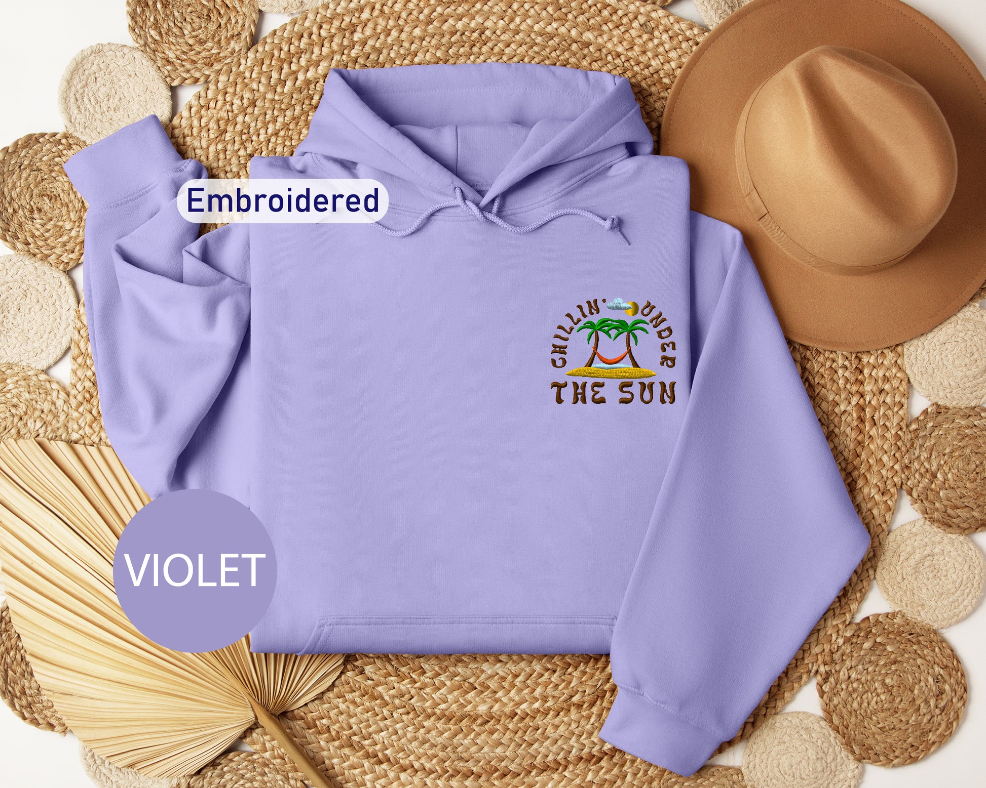 a purple hoodie with a tropical beach design, a brown hat, and a woven straw hat, all arranged on a woven mat.