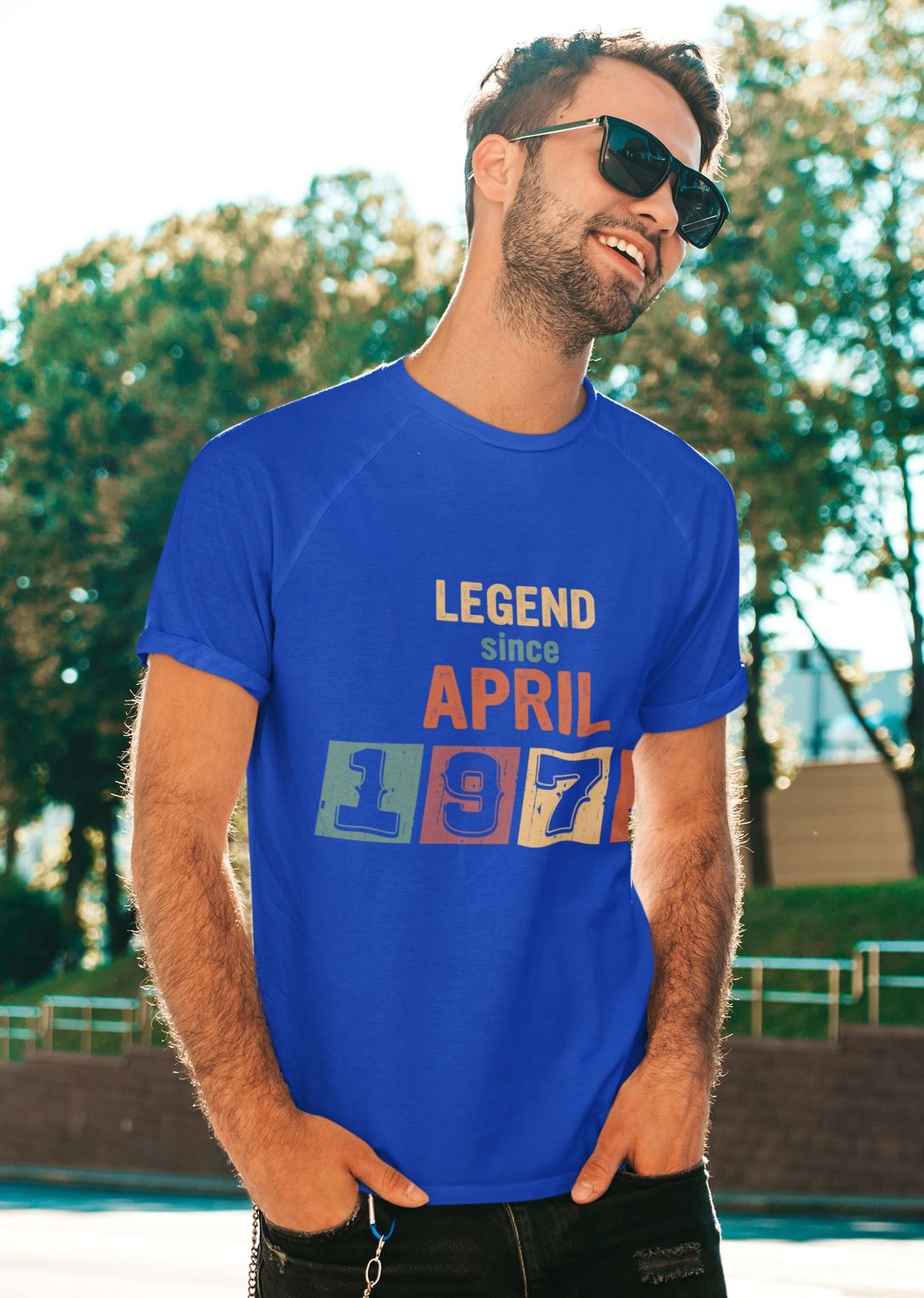 A man wearing a blue t-shirt with the text "LEGEND since APRIL 1877" is smiling and standing outdoors in front of a natural background.