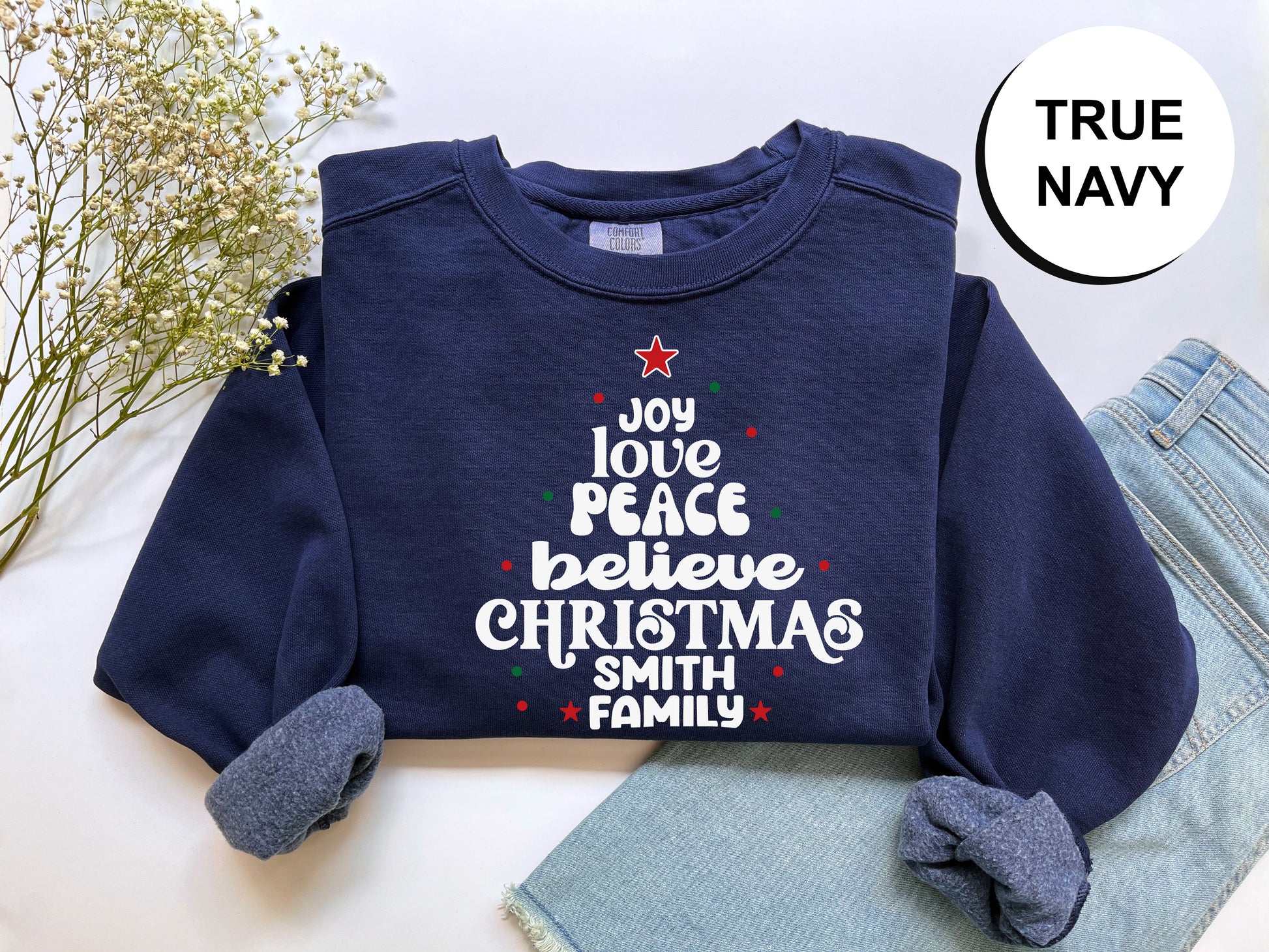 A navy blue sweatshirt with a Christmas tree design and text printed on it, along with a pair of jeans and some flowers.