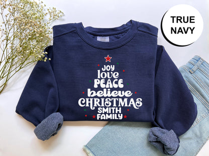 A navy blue sweatshirt with a Christmas tree design and text printed on it, along with a pair of jeans and some flowers.