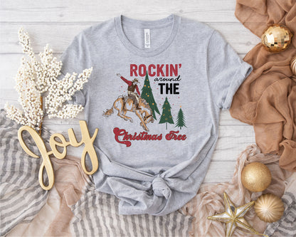 a gray t-shirt with a graphic design of a cowboy on a horse, surrounded by Christmas trees, and the text "ROCKIN' AROUND THE CHRISTMAS Tree". The t-shirt is placed on a wooden surface, surrounded by various Christmas decorations such as ornaments, a star, and a garland.