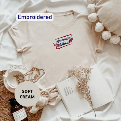 a beige t-shirt with a red and blue embroidered logo, a white bowl, a bottle of lotion, a book, and some dried flowers, all arranged on a white surface.