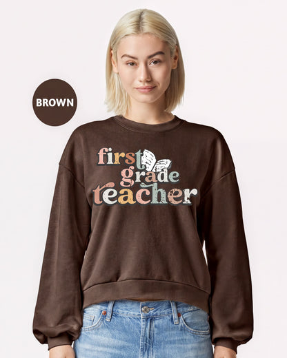 A woman wearing a brown sweatshirt with the text "first grade teacher" printed on it.