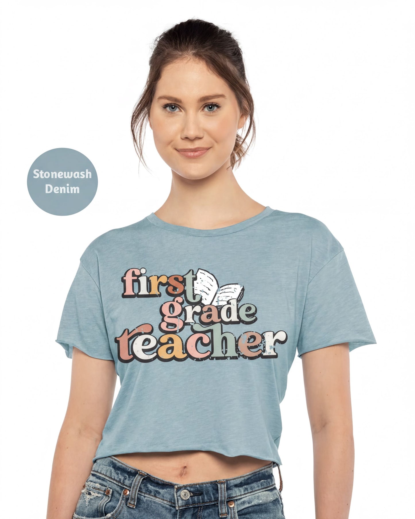 A woman wearing a light blue crop top with the text "first grade teacher" printed on it, smiling at the camera.