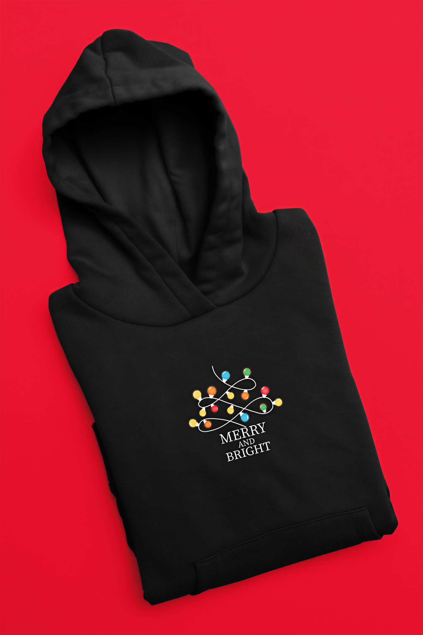 A black hoodie with a festive design and the text "MERRY BRIGHT" printed on it, laid out against a bright red background.