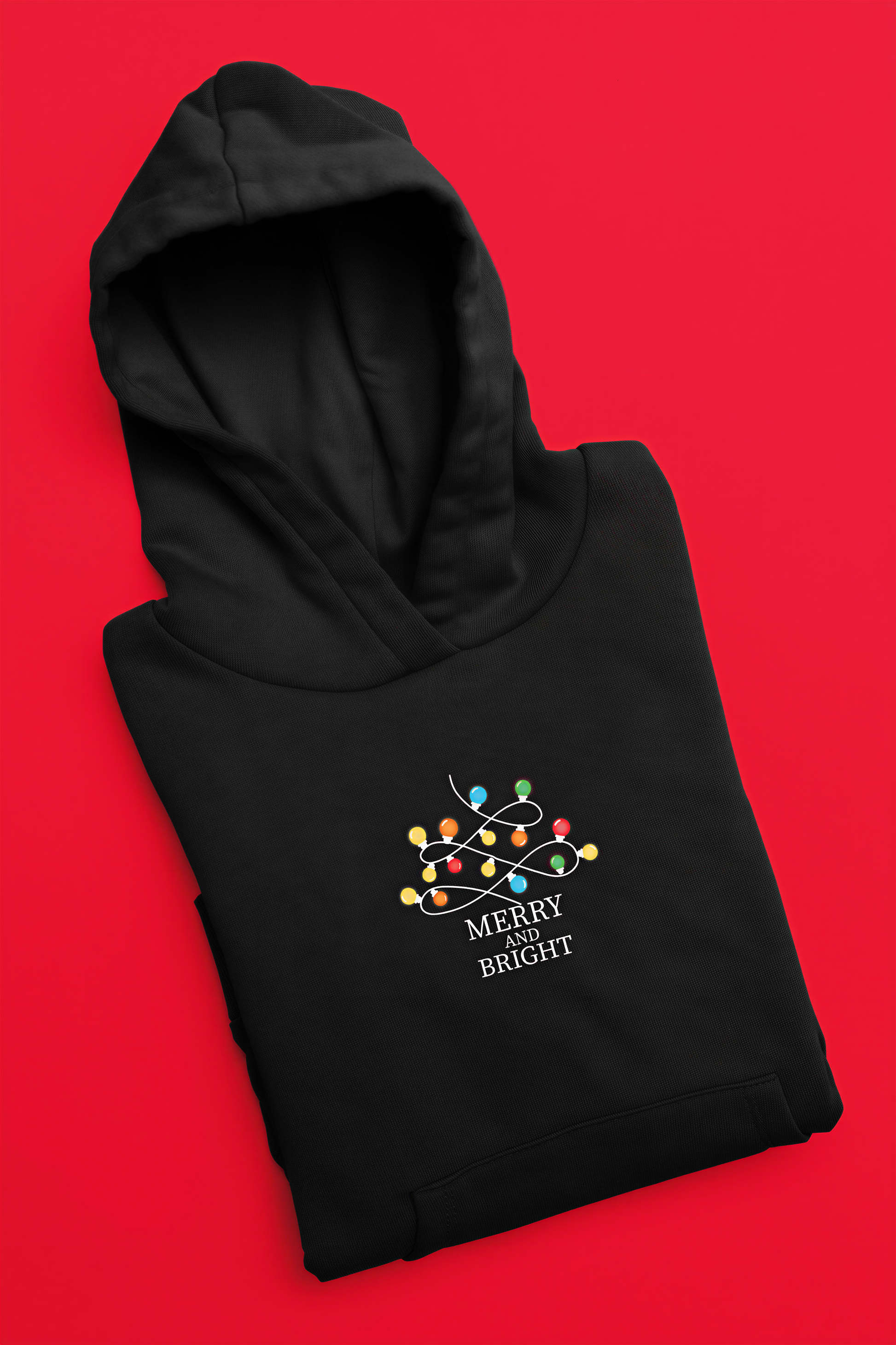 A black hoodie with a festive design and the text "MERRY BRIGHT" printed on it, laid out against a bright red background.