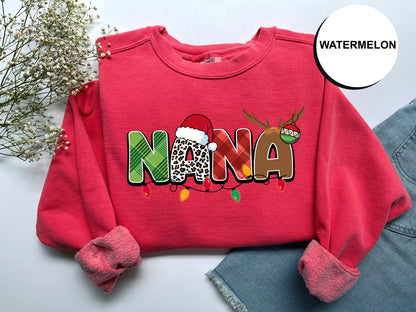 A red sweater with the word "NANA" printed on it, along with a Santa hat and Christmas lights, is displayed on a white surface.