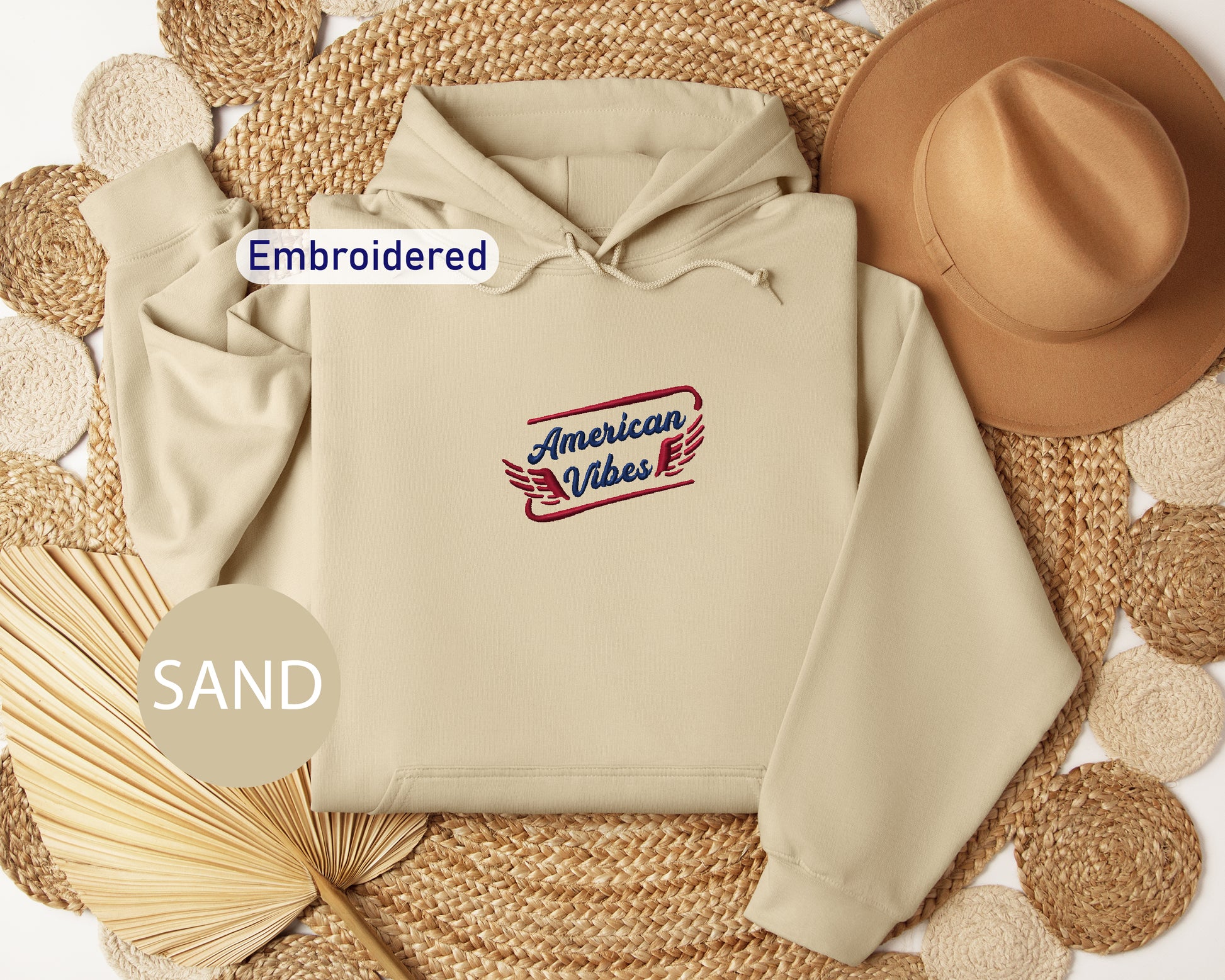 a beige hoodie with an embroidered design, a brown hat, and a fan, all arranged on a woven mat.