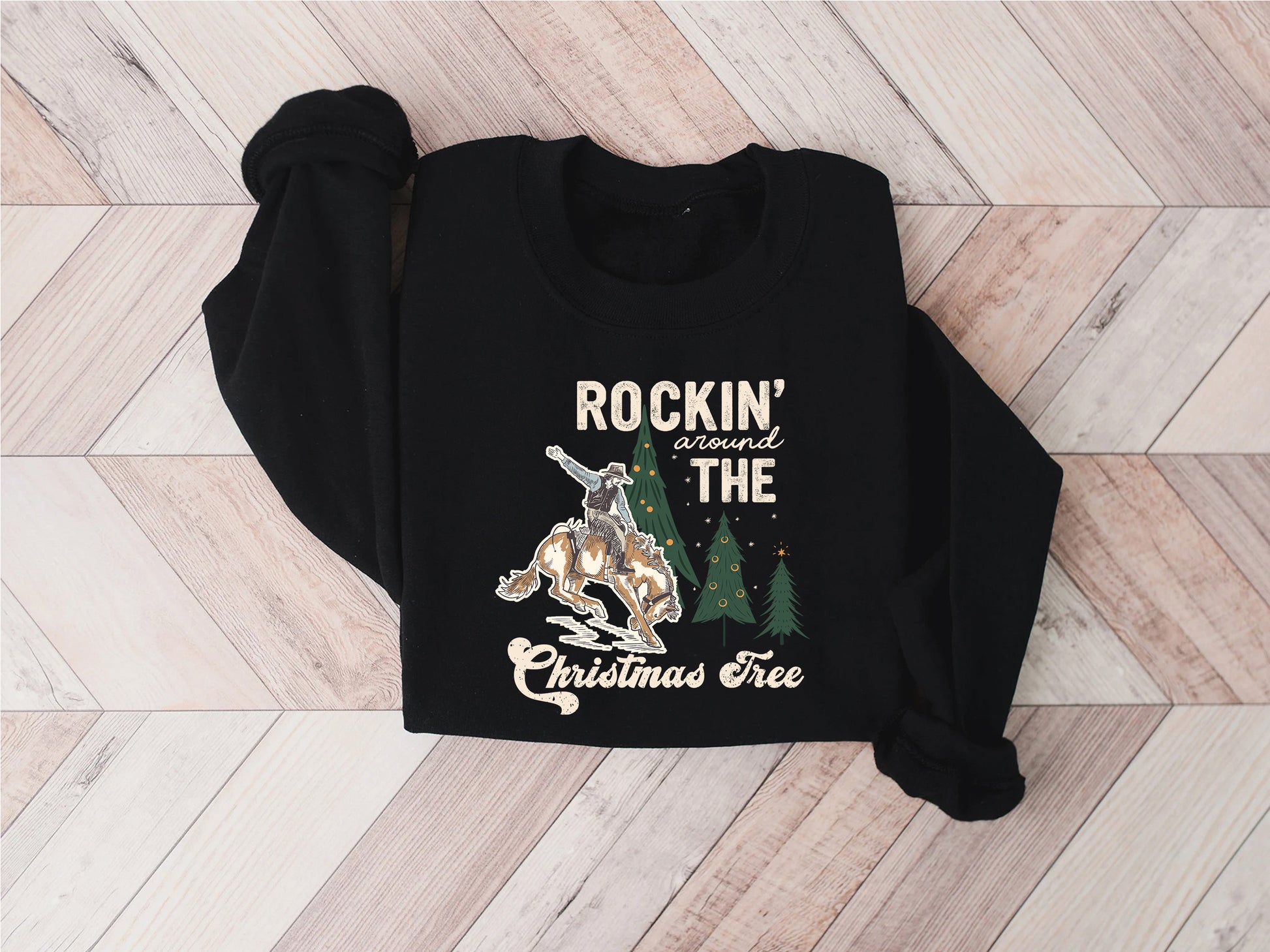 A black sweater with a graphic design on it, featuring a cowboy riding a bucking bronco, a Christmas tree, and the text "ROCKIN' around THE CHRISTMAS Tree".