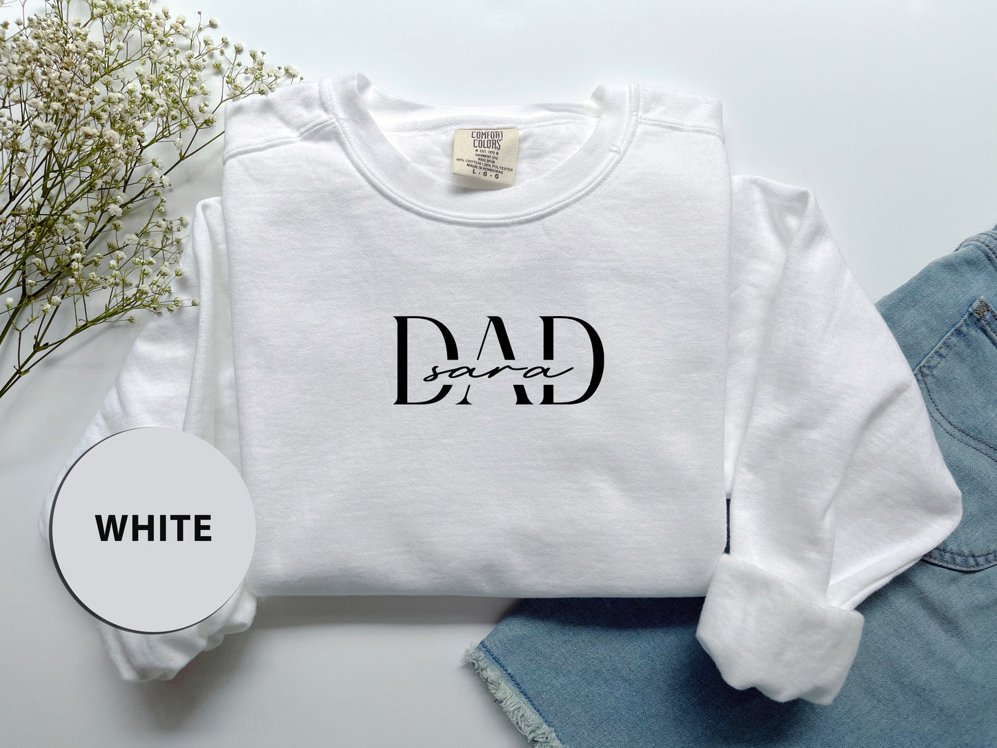 A white long-sleeved shirt with the text "DAD sara" printed on it, along with a pair of blue jeans and some white flowers.