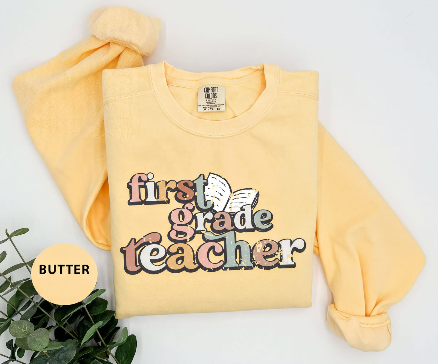 A yellow t-shirt with the text "first grade teacher" printed on it, along with a butterfly graphic. The t-shirt is folded and placed on a white surface, with a plant visible in the background.