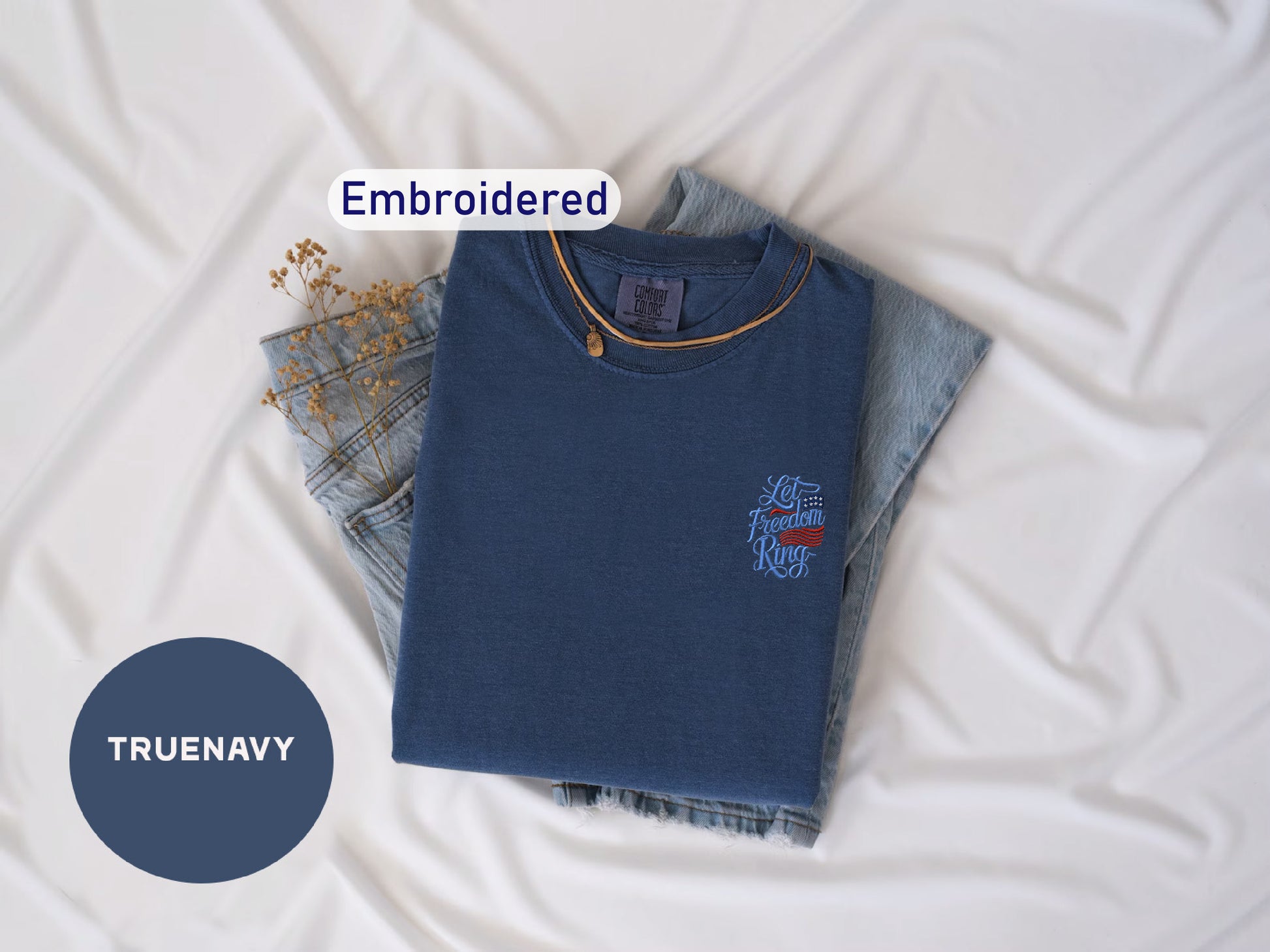a blue t-shirt with a small embroidered design, a pair of denim jeans, and some dried flowers, all laid out on a white surface.