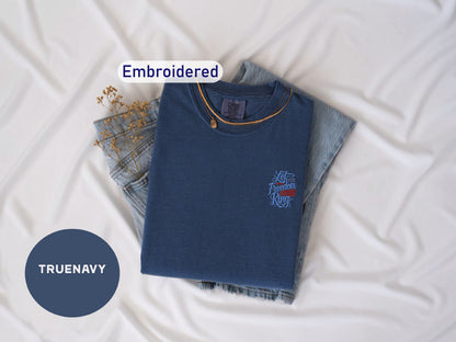 a blue t-shirt with a small embroidered design, a pair of denim jeans, and some dried flowers, all laid out on a white surface.