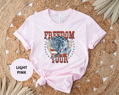 A light pink t-shirt with a graphic design featuring an eagle and the text "FREEDOM TOUR" printed on it, placed on a woven mat with leaves.