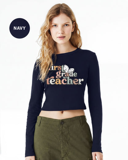 A woman wearing a navy blue crop top with the text "First Grade Teacher" printed on it, standing against a white background.