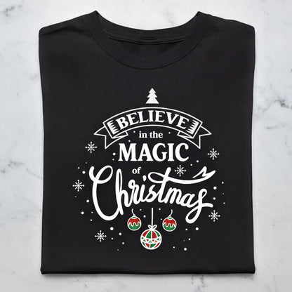 A black t-shirt with a white and red design featuring the text "Believe in the Magic of Christmas" and various Christmas-themed elements such as snowflakes, ornaments, and a small Christmas tree.