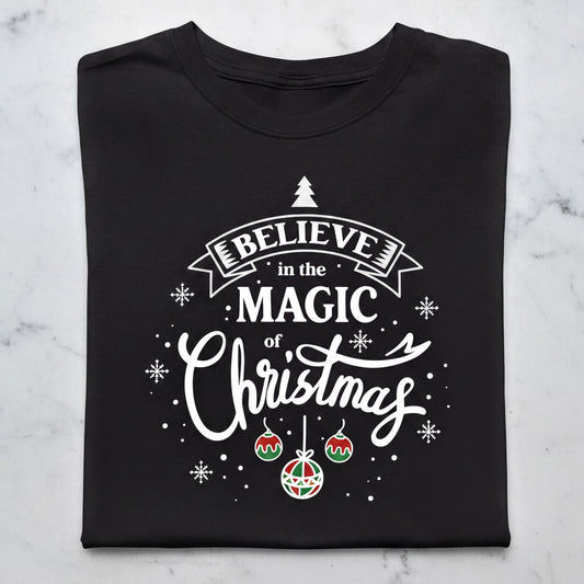 A black t-shirt with a white and red design featuring the text "Believe in the Magic of Christmas" and various Christmas-themed elements such as snowflakes, ornaments, and a small Christmas tree.