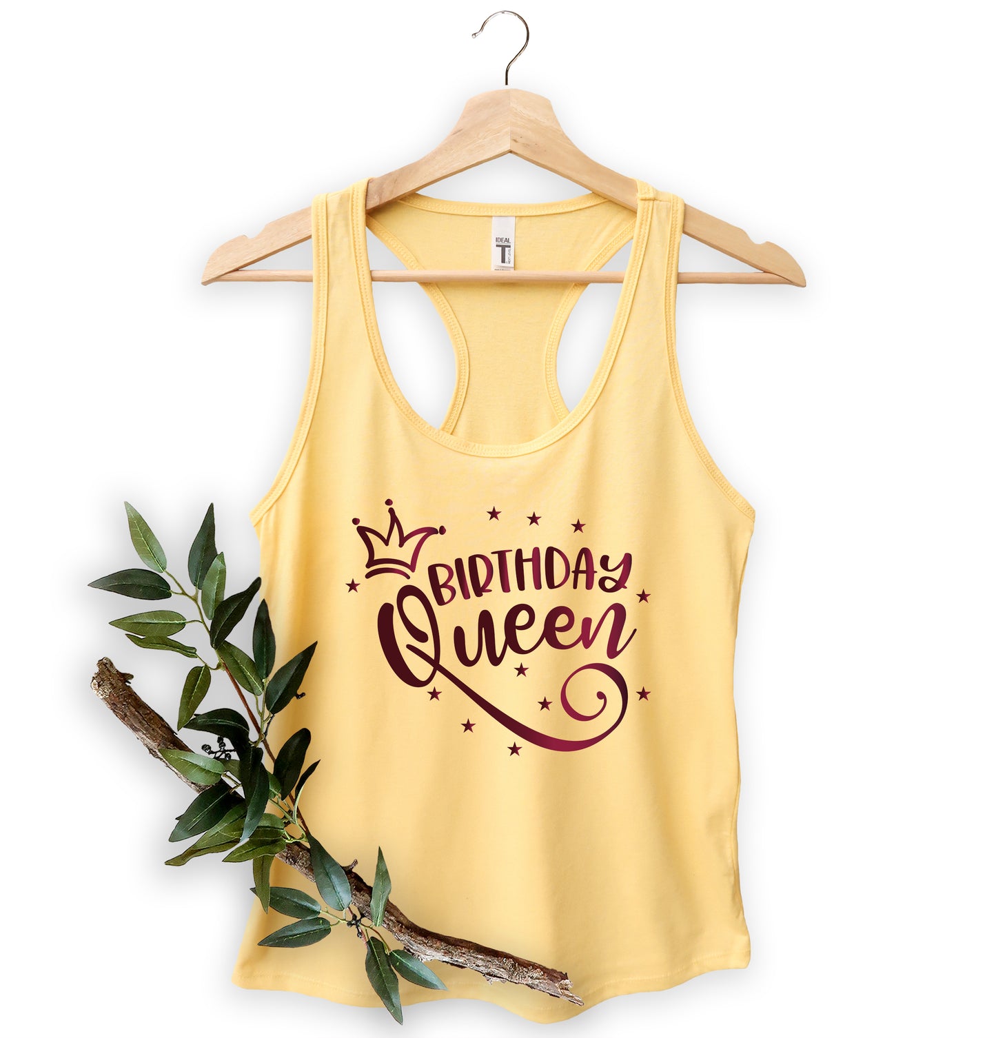 A yellow tank top with the text "BIRTHDAY QUEEN" printed on it, along with a crown and stars. The tank top is hanging on a wooden hanger, and there is a branch with green leaves next to it.