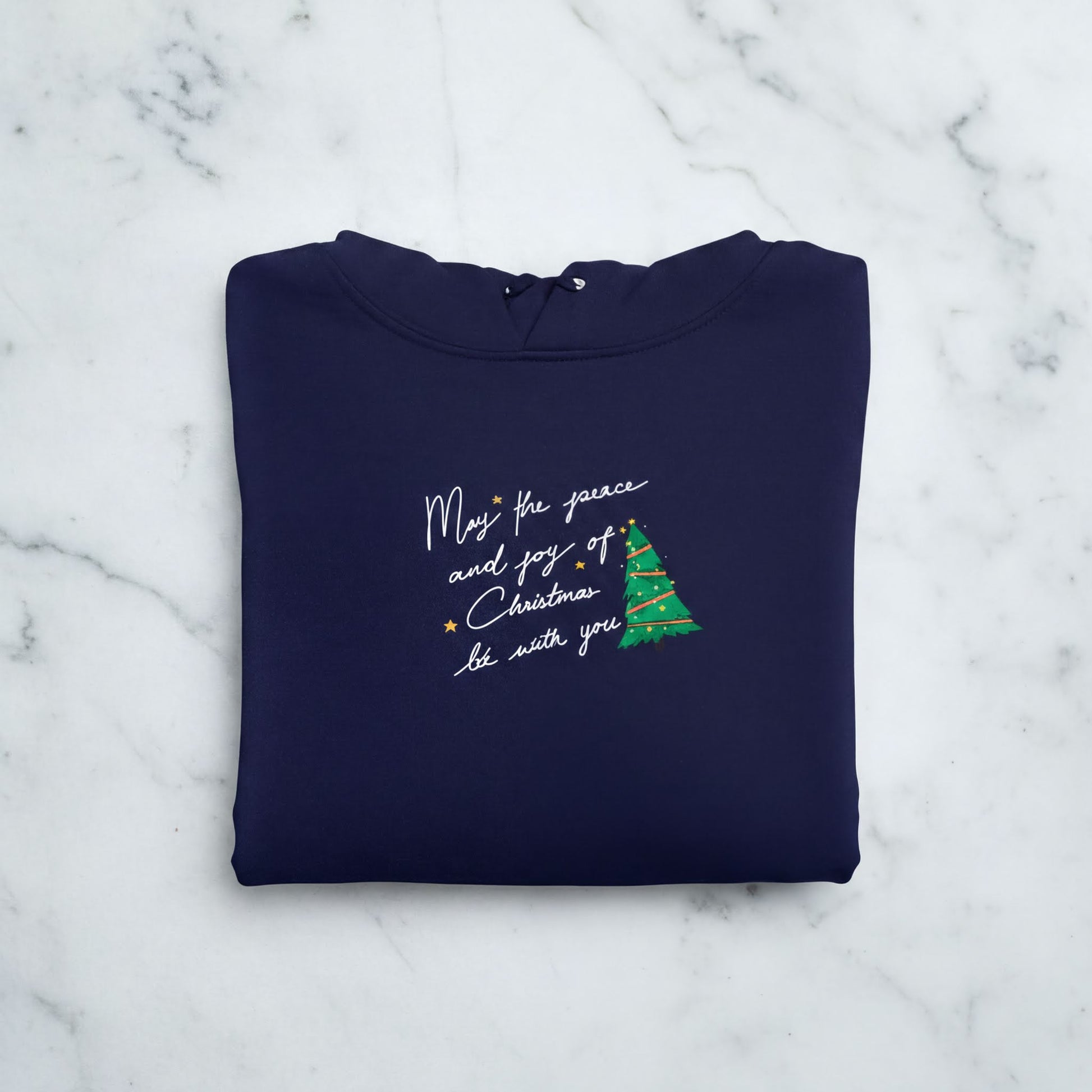 A navy blue hoodie with a Christmas tree design and text printed on it, laid out on a marble surface.