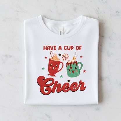 A white t-shirt with a graphic design featuring two colorful mugs and the text "Have a Cup of Cheer" along with stars and other festive elements.