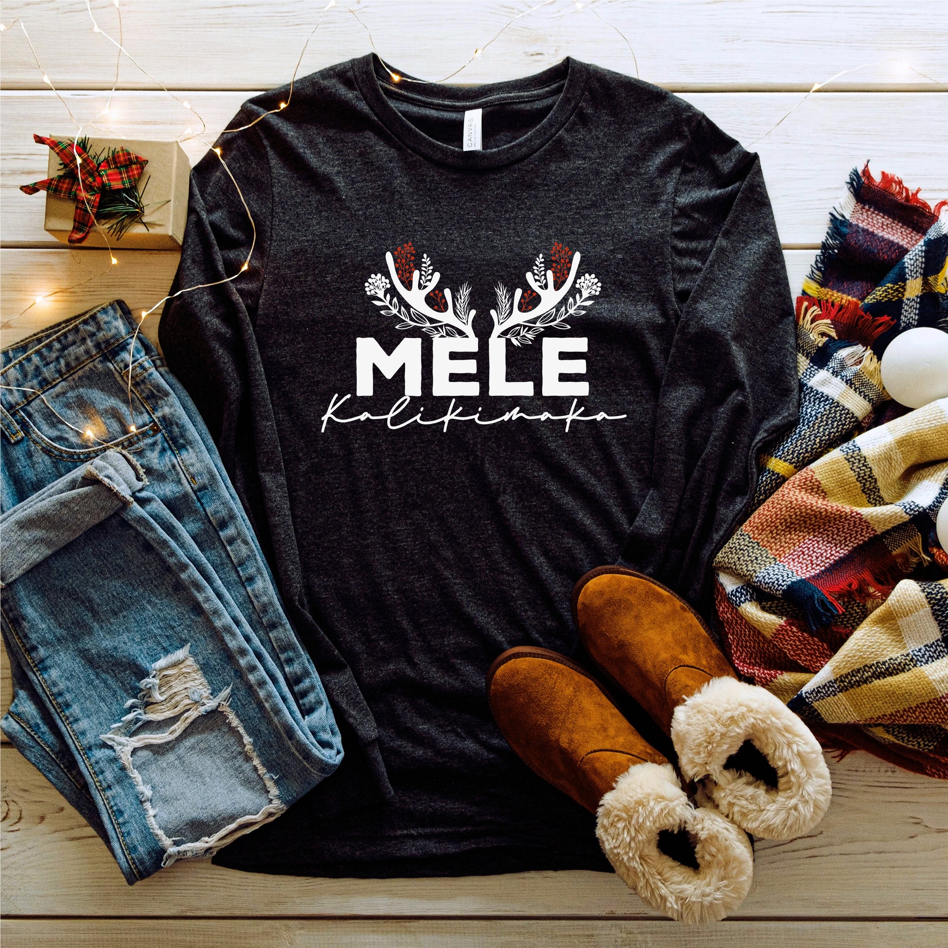A casual outfit consisting of a black long-sleeved shirt with a white text design, distressed blue jeans, brown boots, and a colorful plaid scarf is laid out on a wooden surface, accompanied by a small gift box and twinkling Christmas lights.