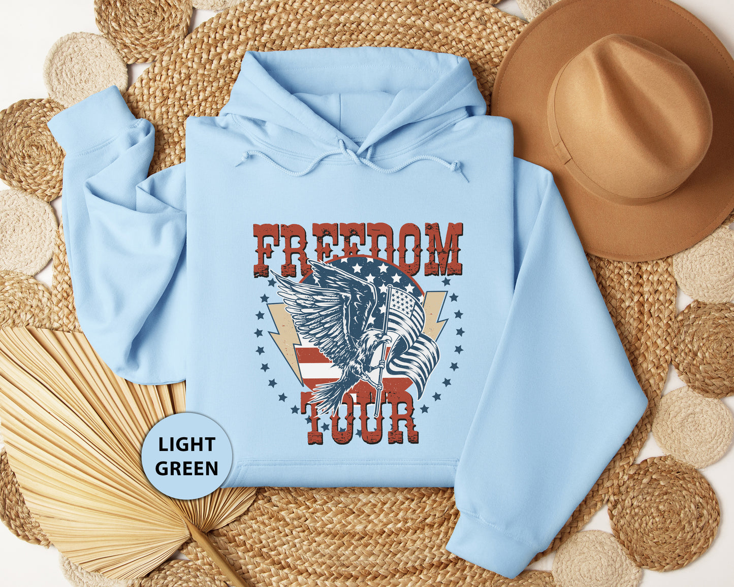 A light blue hoodie with a graphic design featuring a winged eagle and the text "FREEDOM TOUR" is displayed on a woven mat, along with a brown hat and a straw hat.