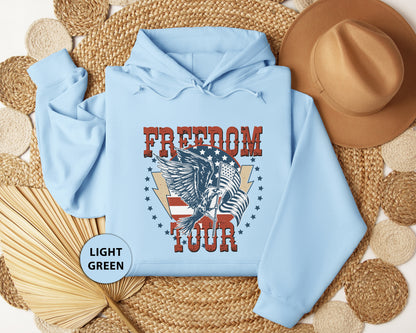 A light blue hoodie with a graphic design featuring a winged eagle and the text "FREEDOM TOUR" is displayed on a woven mat, along with a brown hat and a straw hat.