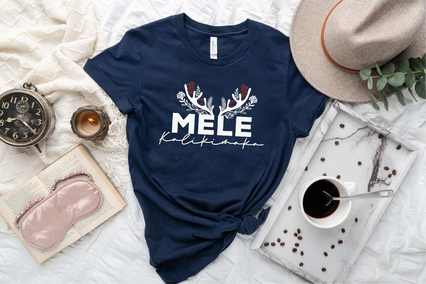 A navy blue t-shirt with the text "MELE KOLIKINAS" printed on it, along with a hat, a book, a cup of coffee, and some other items arranged on a bed.