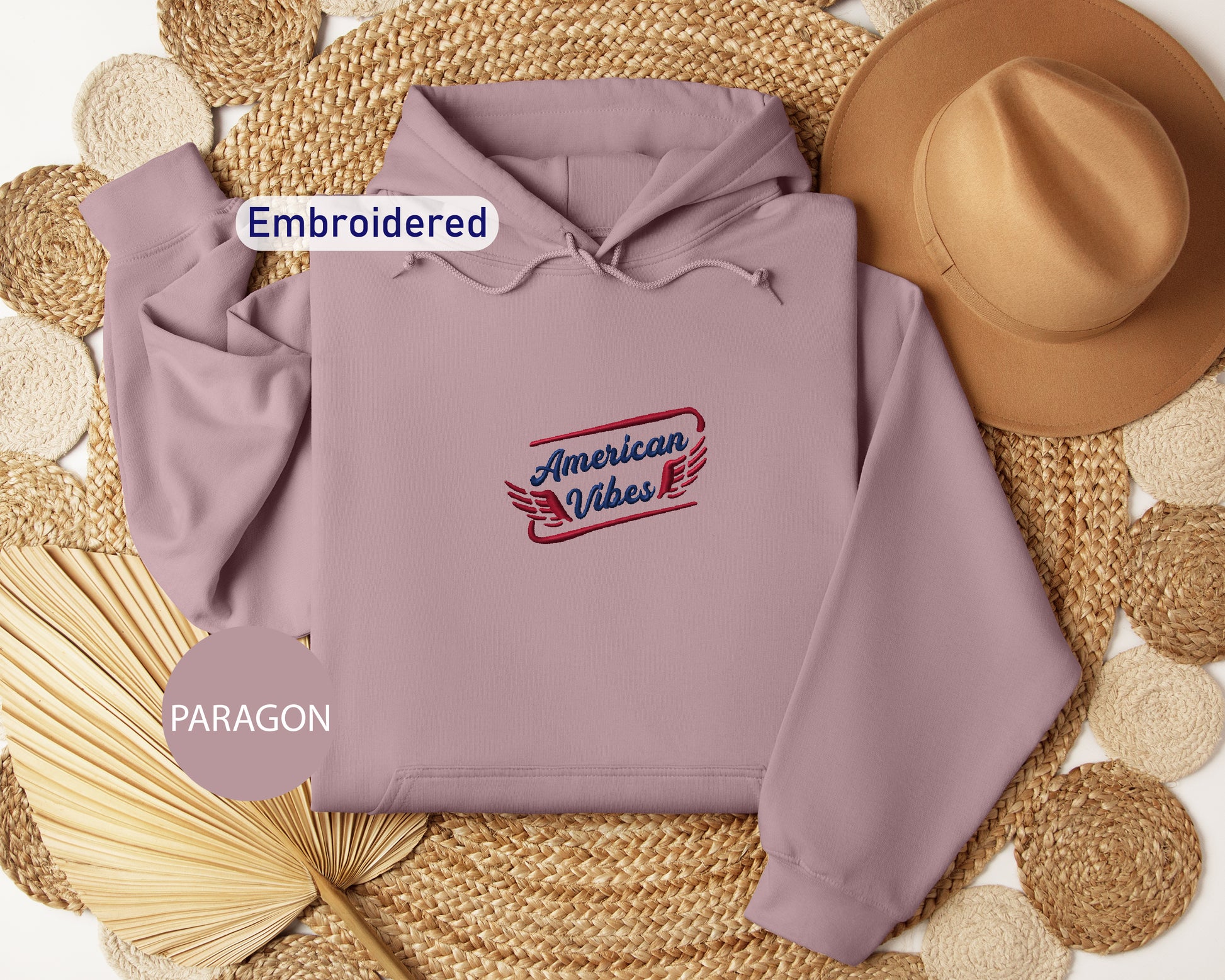 a gray hoodie with a red and blue embroidered logo, a brown hat, and a fan, all arranged on a woven mat.