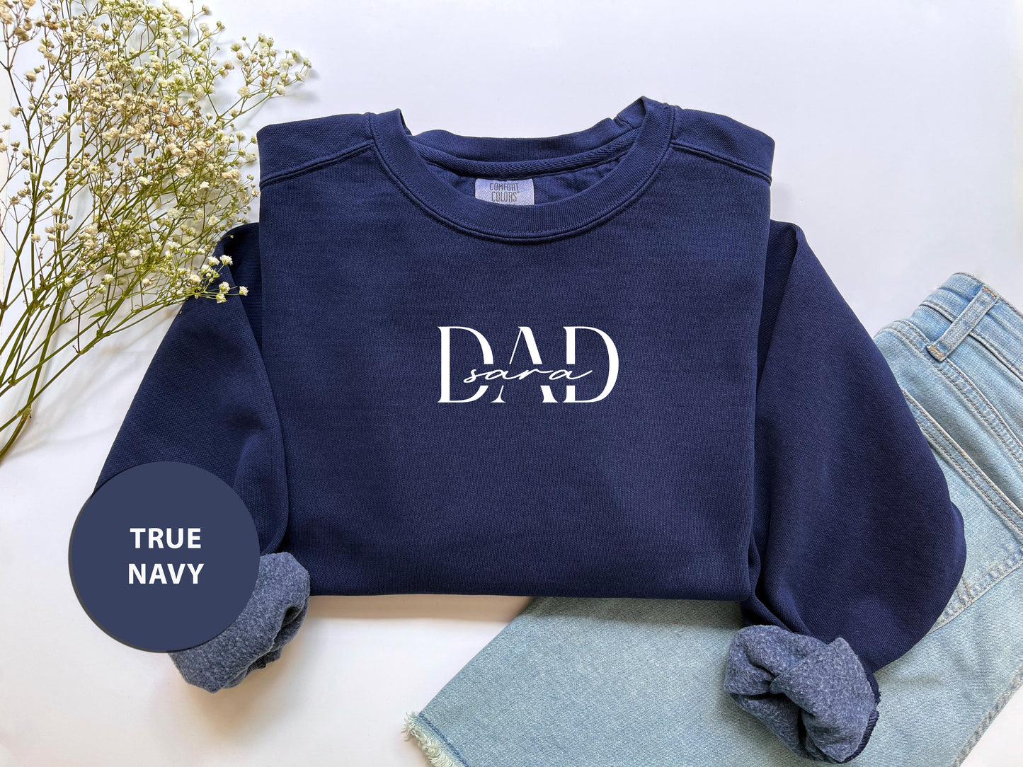 A navy blue sweatshirt with the text "DAD" printed on it, along with a pair of blue jeans and a floral arrangement, all laid out on a white background.