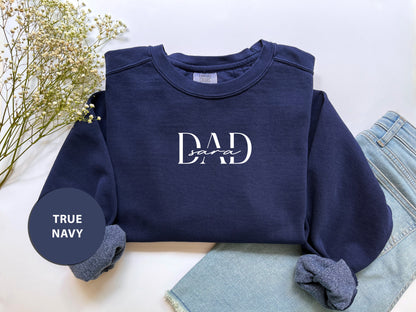 A navy blue sweatshirt with the text "DAD" printed on it, along with a pair of blue jeans and a floral arrangement, all laid out on a white background.