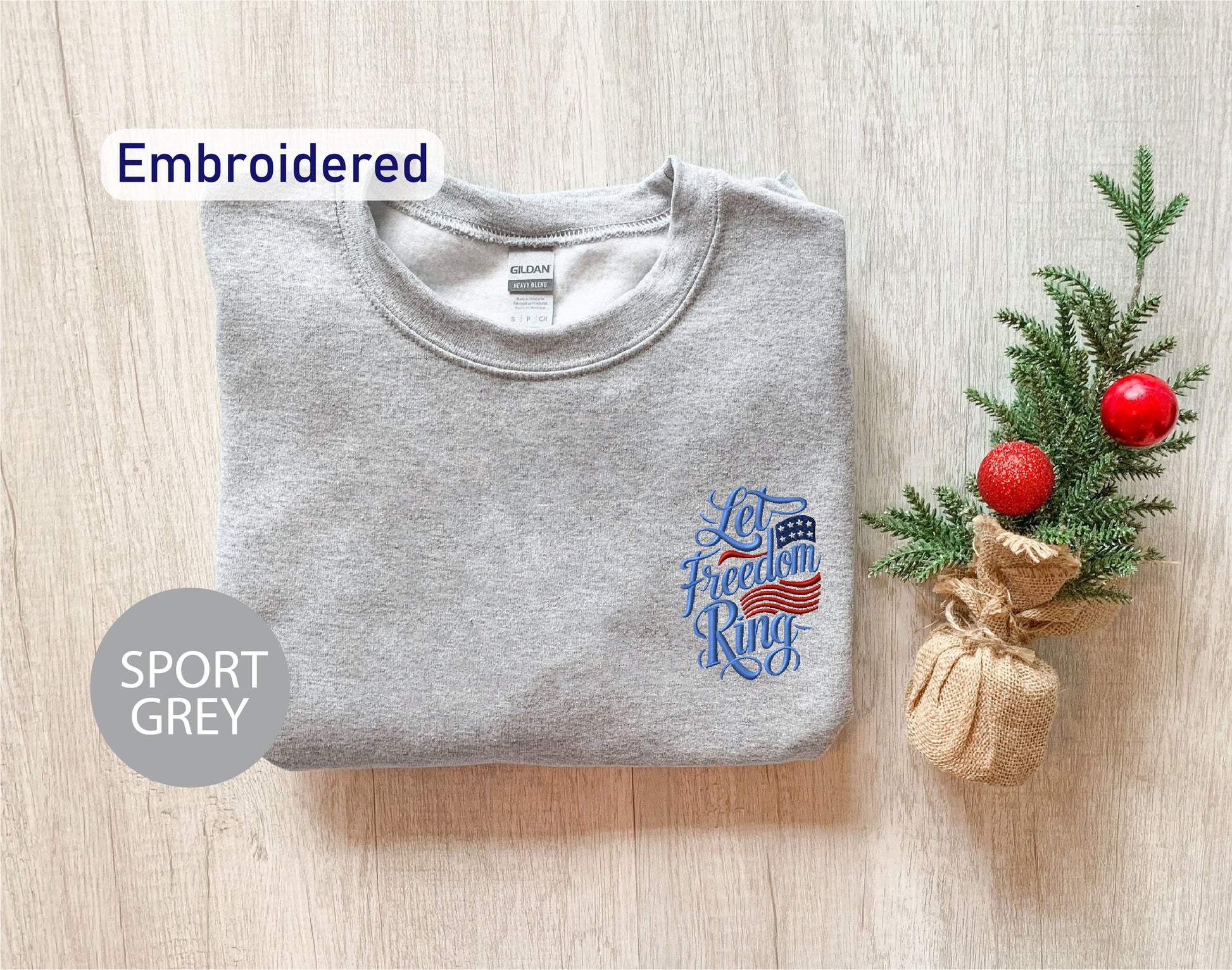 a grey sports sweatshirt with an embroidered design on the left chest area, along with a small Christmas tree and a burlap sack on the right side.