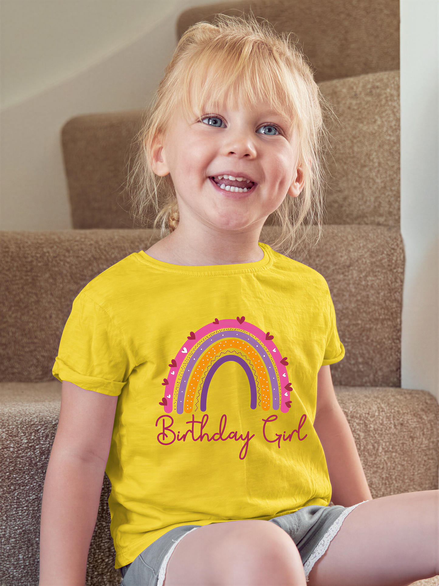 A young girl with blonde hair is sitting on a carpeted staircase, wearing a yellow t-shirt with a rainbow graphic and the text "Birthday Girl".