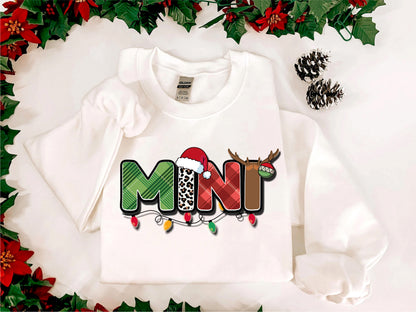 A white t-shirt with a colorful graphic design featuring the word "MINT" in a playful font, surrounded by holiday-themed elements such as pine cones and poinsettias.
