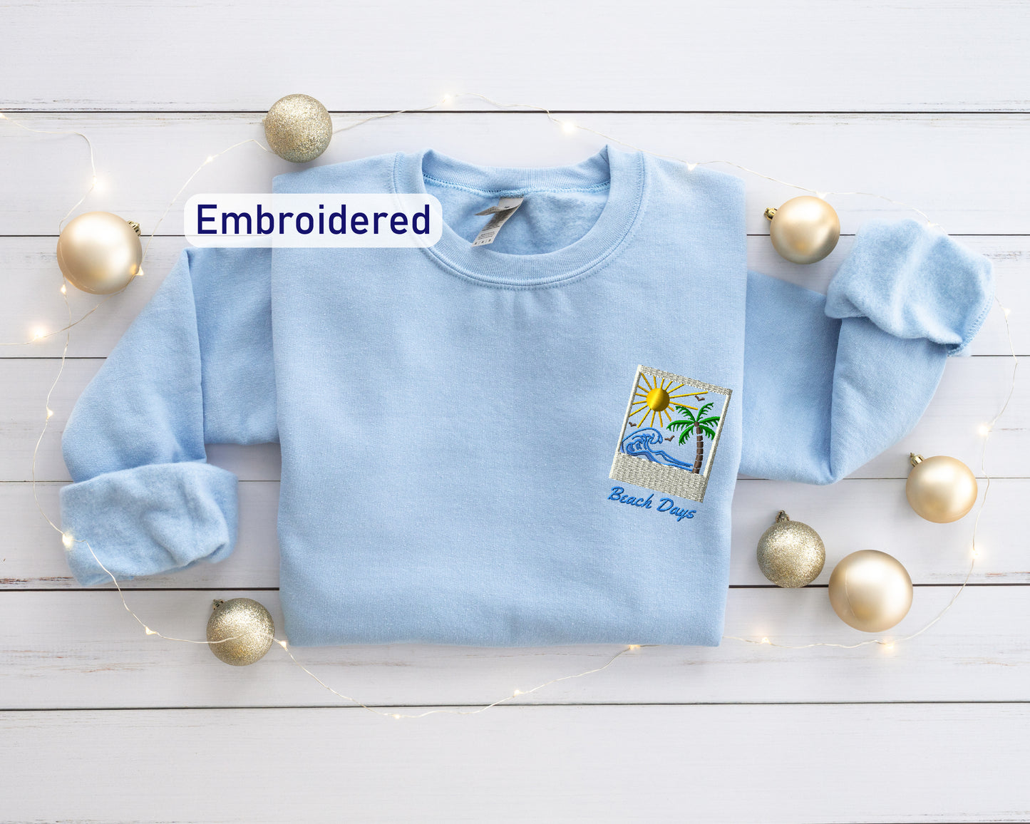a light blue t-shirt with a beach-themed embroidered patch, surrounded by gold ornaments and a string of lights on a white wooden surface.