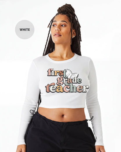 A woman with braids wearing a white crop top with text that says "first grade teacher" printed on it.