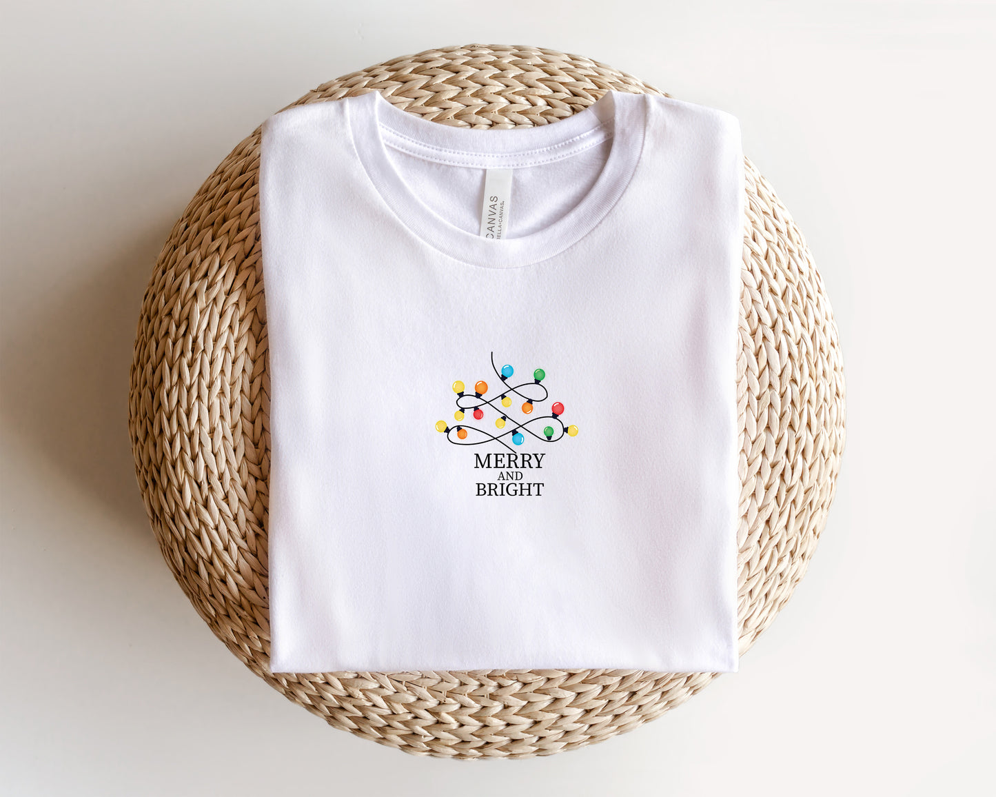 A white t-shirt with a graphic design of colorful Christmas lights and the text "MERRY on BRIGHT" printed on it, laid out on a woven basket.