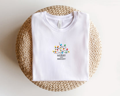 A white t-shirt with a graphic design of colorful Christmas lights and the text "MERRY on BRIGHT" printed on it, laid out on a woven basket.