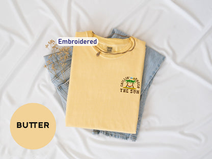 a yellow t-shirt with a graphic design, a pair of blue jeans, and a small plant, all laid out on a white background.