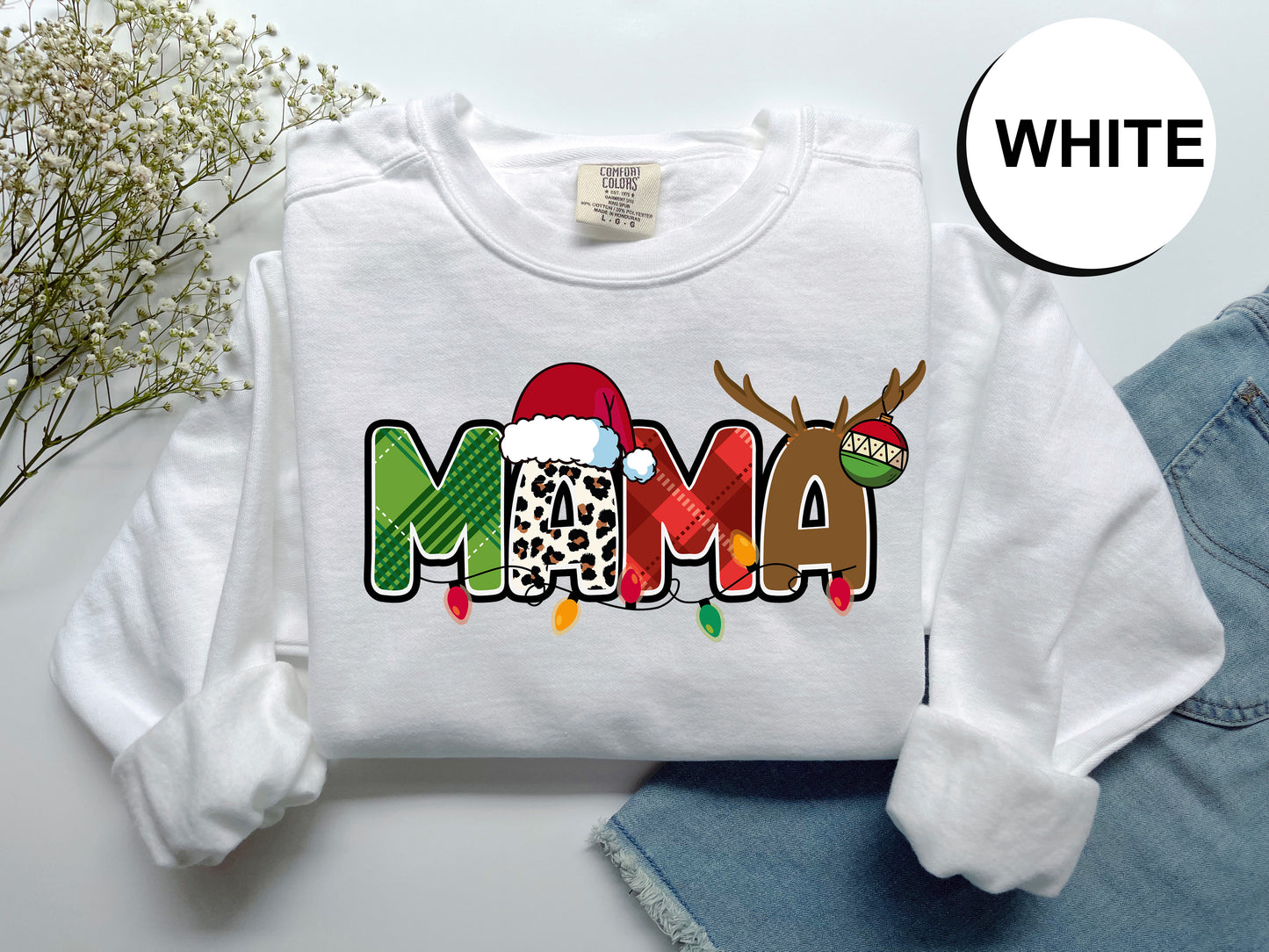 A white t-shirt with a colorful graphic design featuring the word "MAMA" in a playful font, along with a Santa hat and antlers. The t-shirt is displayed on a white background, and there are some decorative elements like a plant and a pair of jeans visible in the image.