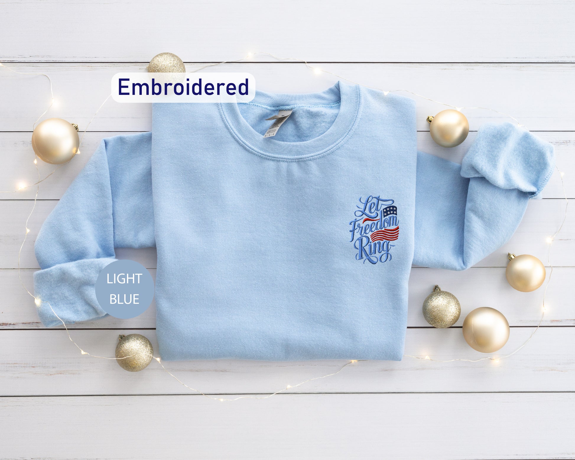 a light blue long-sleeved shirt with an embroidered design on the left side, placed on a white wooden surface. Surrounding the shirt are gold ornaments, and there is a string of lights visible in the background.