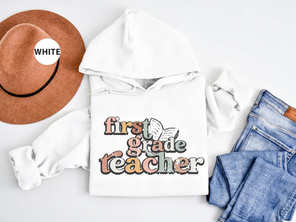 A white hoodie with the text "first grade teacher" printed on it, along with a brown hat and a pair of blue jeans, all laid out on a white background.