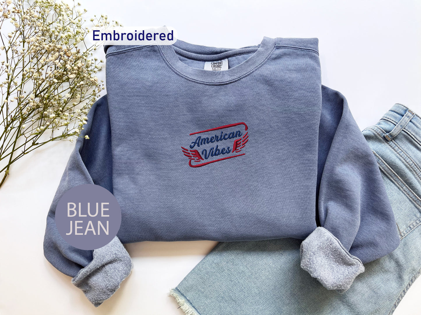 a blue sweatshirt with an embroidered design, a pair of blue jeans, and some flowers on a white background.