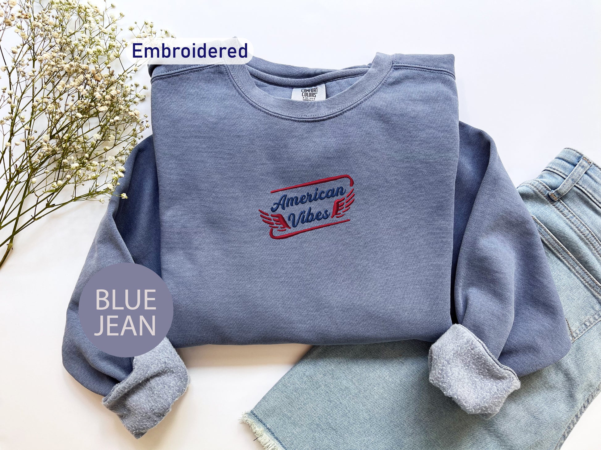 a blue sweatshirt with an embroidered design, a pair of blue jeans, and some flowers on a white background.