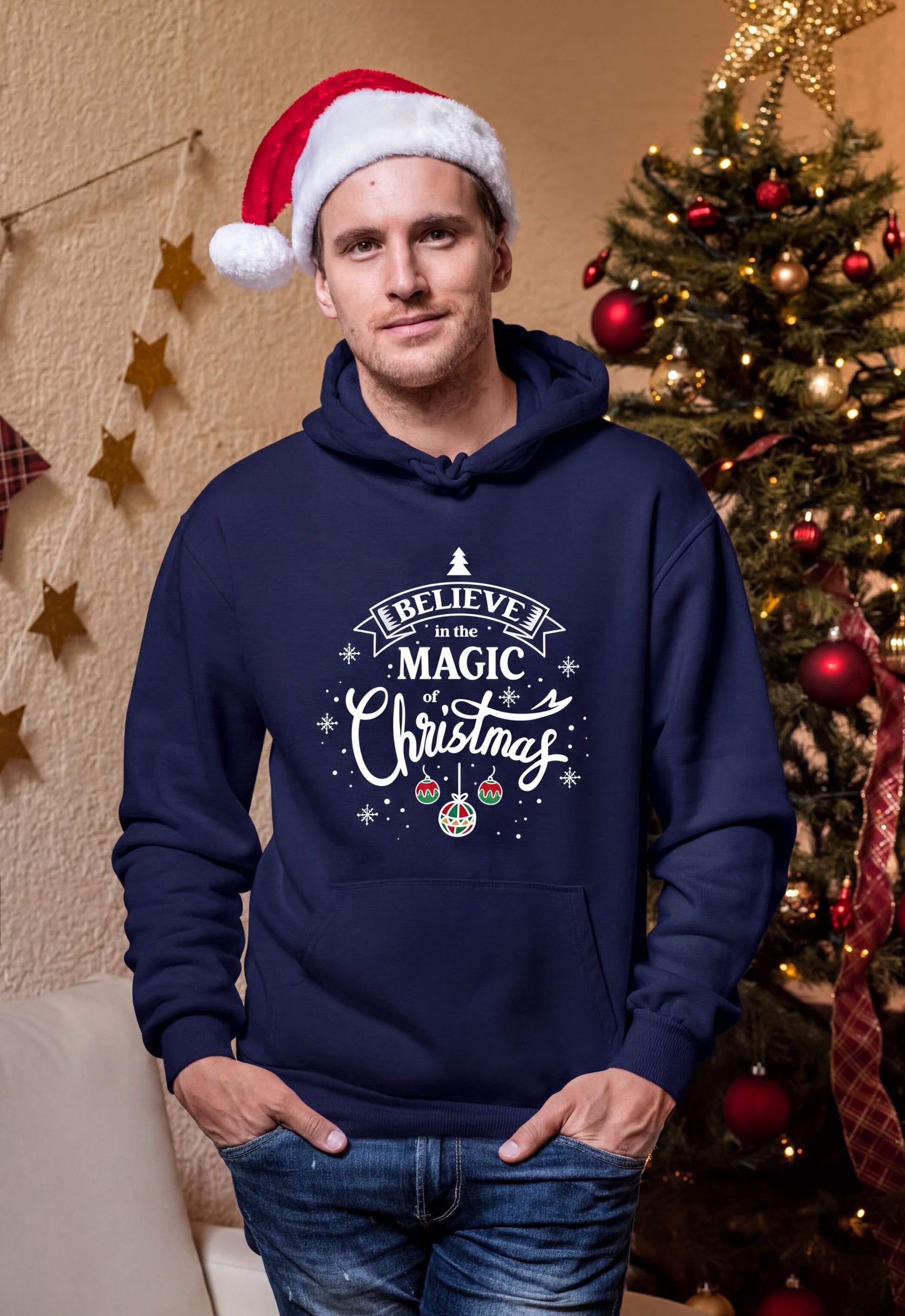 A man wearing a navy blue hoodie with a Christmas-themed design stands in front of a decorated Christmas tree.