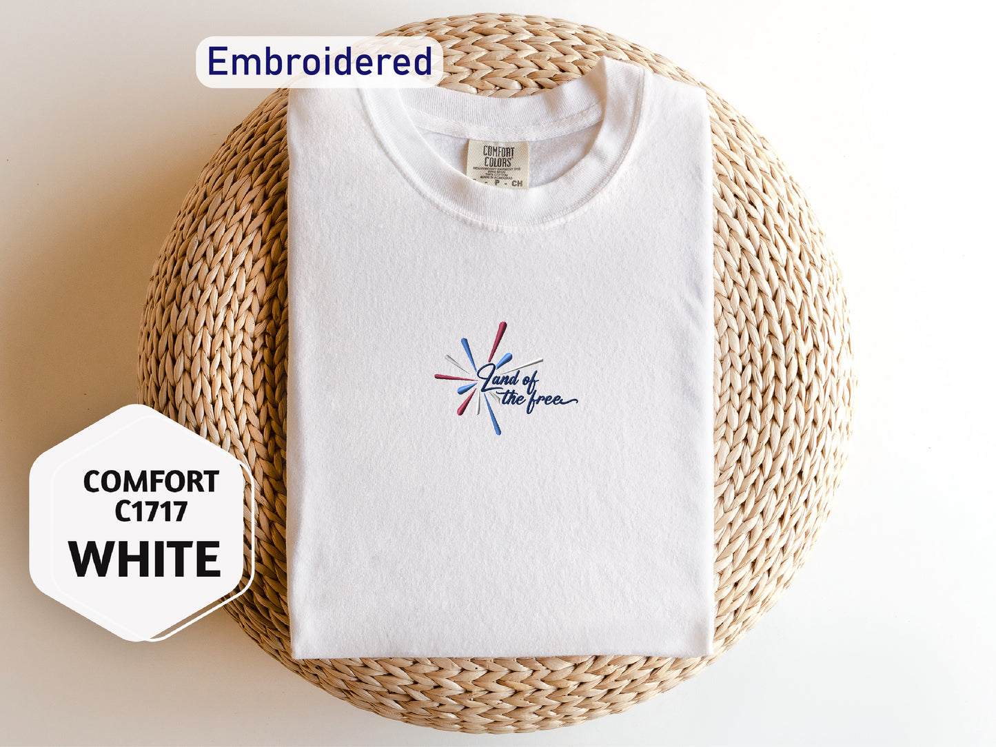 a white t-shirt with a colorful design, laid out on a woven basket or mat. The t-shirt has a label on the collar, and there is a hexagonal sticker next to it with text.